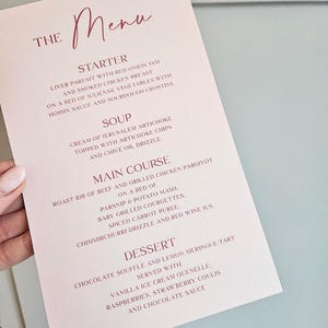 May include: A pink menu card with a handwritten title "THE Menu" in a dark pink colour. The menu features a starter, soup, main course, and dessert. The menu items are listed in a simple, elegant font.