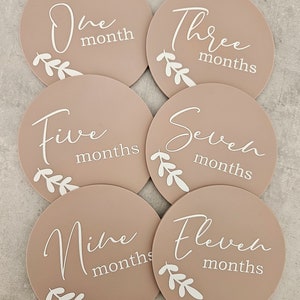 May include: Six round, beige baby milestone cards with white script and floral designs. The cards read "One month", "Three months", "Five months", "Seven months", "Nine months", and "Eleven months".