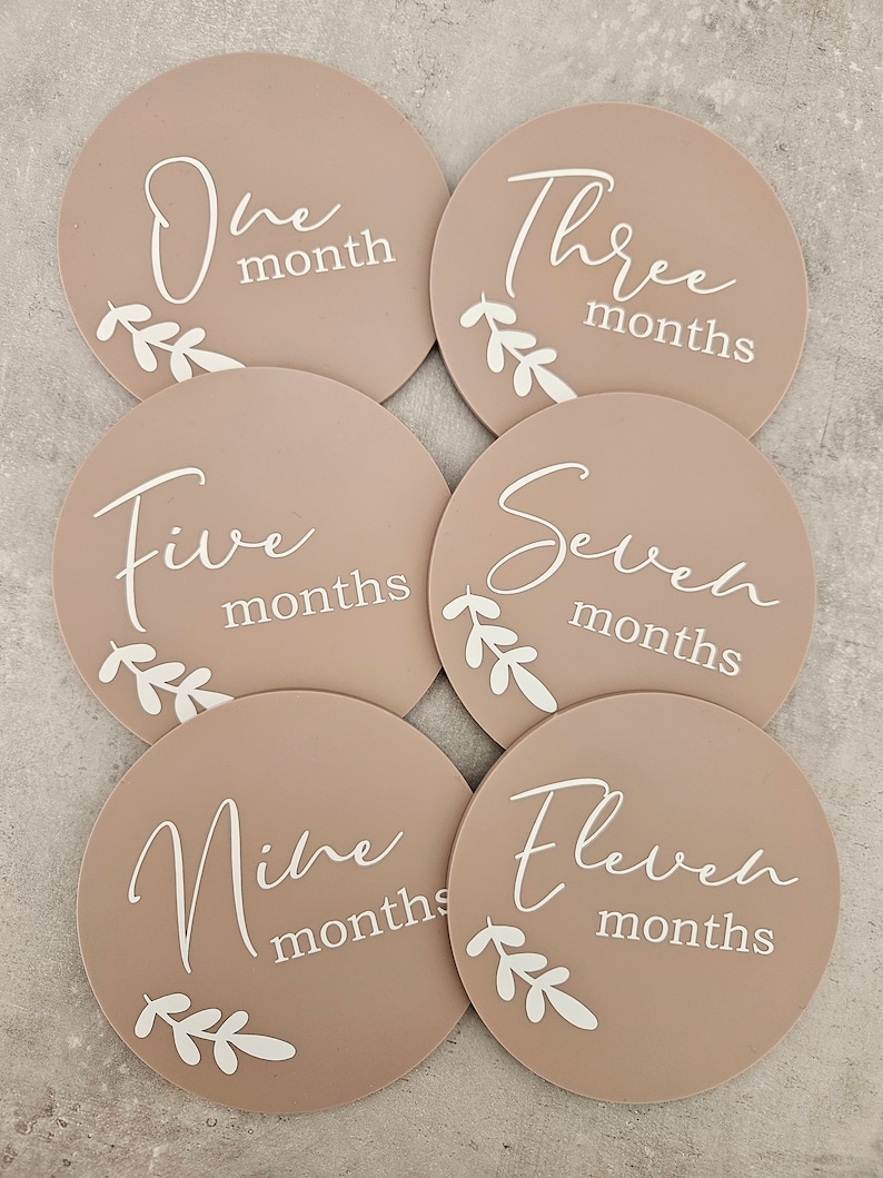 May include: Six round, beige baby milestone cards with white script and floral designs. The cards read "One month", "Three months", "Five months", "Seven months", "Nine months", and "Eleven months".