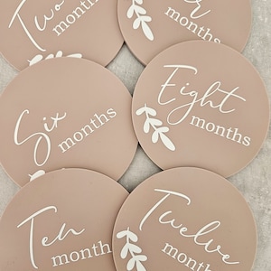 May include: Six round beige baby milestone cards with white text and leaves. The cards read "Two months", "Four months", "Six months", "Eight months", "Ten month", and "Twelve months".