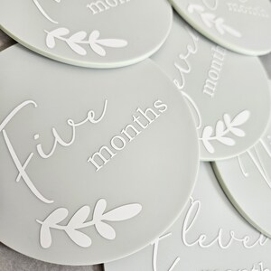 May include: A set of gray baby milestone discs with white text and leaf designs. The discs feature the numbers one through twelve with the word "months" written below each number.
