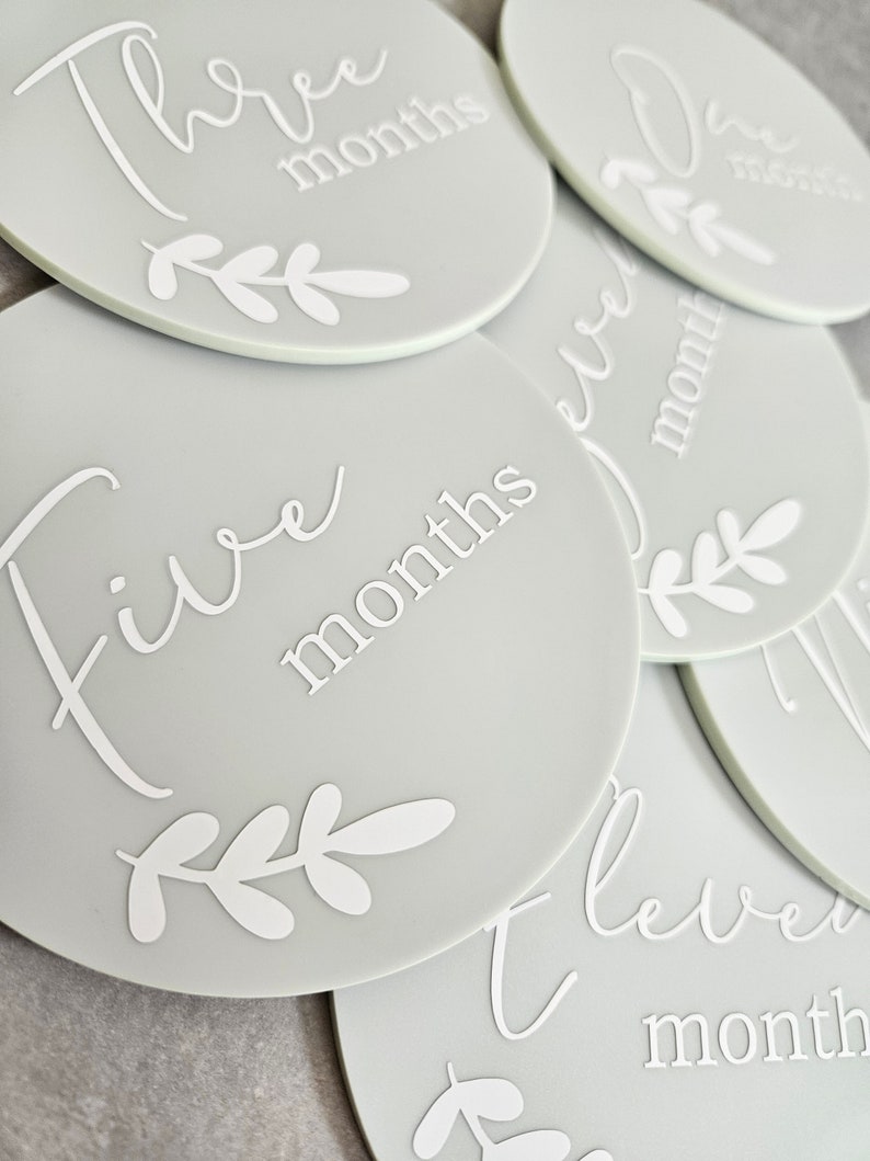 May include: A set of gray baby milestone discs with white text and leaf designs. The discs feature the numbers one through twelve with the word "months" written below each number.