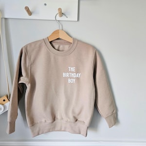 May include: A beige sweatshirt with the text "THE BIRTHDAY BOY" printed in white on the front.