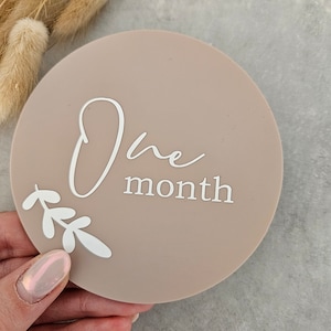 May include: A round, light brown milestone marker with the text "One month" in white script font. The marker has a white floral design around the bottom.