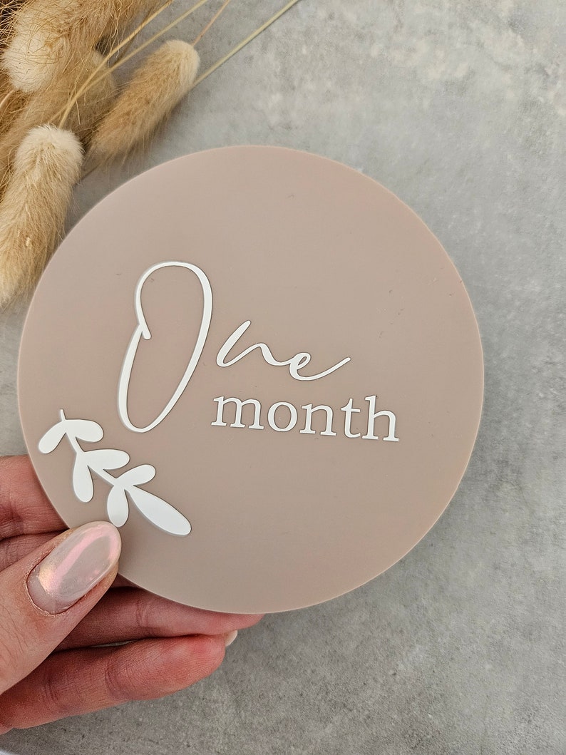 May include: A round, light brown milestone marker with the text "One month" in white script font. The marker has a white floral design around the bottom.
