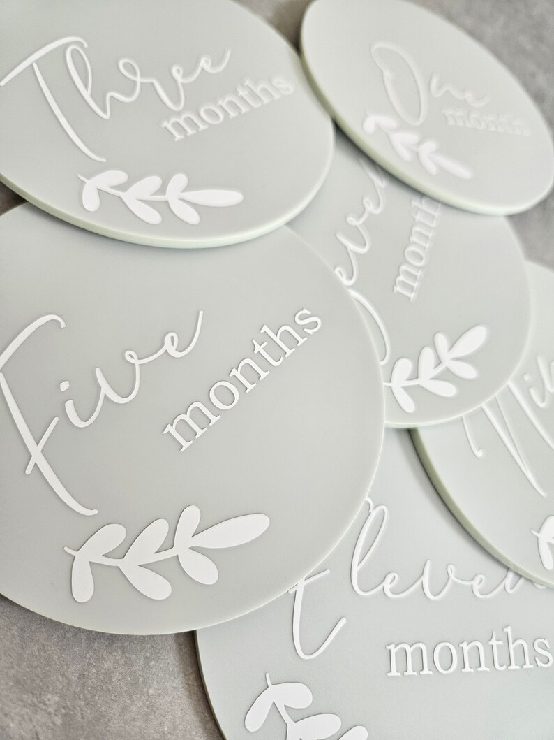 May include: A set of ten gray baby milestone discs with white text and a leafy design. The discs feature the numbers one through ten with the word "months" written below each number.