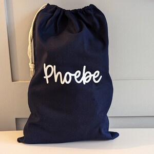 May include: A navy blue drawstring bag, featuring the name "Phoebe" in white script. It has a natural-coloured drawstring closure. The bag is made of a soft fabric and is standing upright.