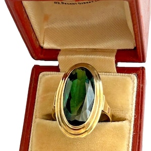 Könnte beinhalten: Ein Goldring mit einem großen, oval geschliffenen, dunkelgrünen Edelstein in einer glatten Fassung, präsentiert in einer roten, aufklappbaren Schmuckschatulle mit cremefarbenem Samtinnenfutter und dem Aufdruck 112 REGENT STREET im Deckel.