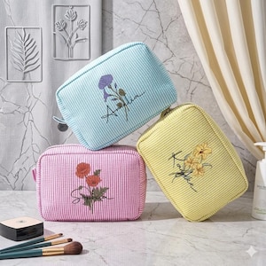 May include: Three striped cosmetic bags in pastel colors: light blue, pink, and yellow. Each bag features embroidered floral designs and names: Amelia, Sophia, and Kaylee. Makeup brushes and a compact are visible.