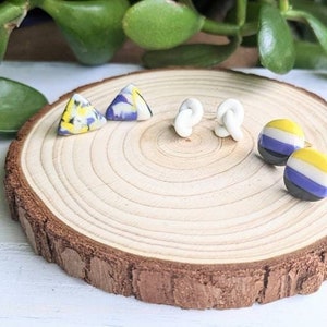 May include: A collection of handmade earrings displayed on a wooden slice. The earrings include triangular, knot-shaped, and round stud designs in various colors, including yellow, blue, white, and black. Green foliage is visible in the background.