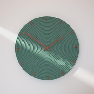 Könnte beinhalten: Eine runde, grüne Wanduhr mit einem strukturierten Design aus konzentrischen Kreisen. Die Uhr hat orange-rote Zeiger und Stundenmarkierungen. Auf dem Zifferblatt steht unten das Wort "wonderfactory".