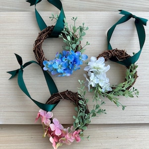 May include: Three small wreaths decorated with faux flowers and greenery. The wreaths are tied with green ribbon and are perfect for a spring or summer decor.