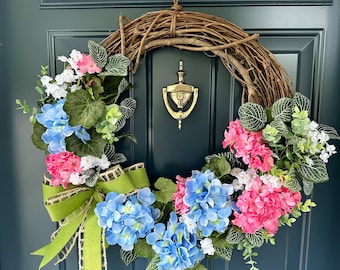 May include: A spring wreath with a brown vine base, decorated with pink and blue hydrangeas, white flowers, green leaves, and a green bow.