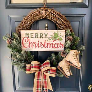 May include: A Christmas wreath with a wooden sign that says "Merry Christmas" in red and green lettering. The wreath is decorated with pine branches, a red and black plaid bow, and two white ice skates.