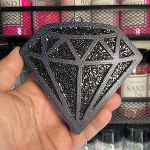 May include: A diamond-shaped ornament with a glittery black center and a dark gray outline. The ornament has a silver loop for hanging. The background shows jars of glitter in various colors.
