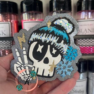 May include: A decorative skull-shaped item with a silver glitter border. The skull has black eyes, a white face, and a blue and silver glitter hair bun. A gold lightning bolt and a skeleton hand with a peace sign are also featured.