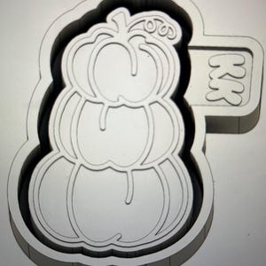 Può includere: Tagliapasta in plastica bianca a forma di zucca impilata, con le lettere "KK" su una piccola estensione rettangolare. Il tagliapasta ha un bordo spesso, progettato per la cottura.