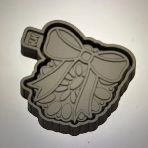 Op de afbeelding: Een grijze plastic koekjesvormer in de vorm van een krans met een strik erbovenop. De koekjesvormer heeft een getextureerd oppervlak en het woord "KK" is aan de zijkant gegraveerd.