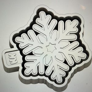 May include: A light gray snowflake-shaped cookie cutter with a detailed design. The cutter has a rectangular handle with the letters "KK" engraved on it. The snowflake has a star in the center and intricate cut-out patterns.