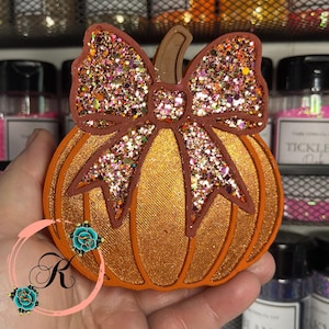 May include: A decorative pumpkin with a glittery bow. The pumpkin is orange with brown outlines and a textured surface. The bow is brown with multi-colored glitter. The pumpkin is held in a hand.