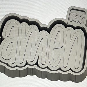 May include: A gray, 3D-rendered design featuring the word "amen" in a rounded, stylized font. The design includes a rectangular tag with the letters "KK". The overall shape is a rounded rectangle.