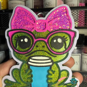 May include: A glittery green frog-shaped keychain with a pink bow and glasses. The frog is holding a blue coffee cup. The keychain has a white outline and a small loop for attaching to keys or a bag.