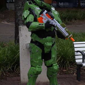 May include: A green and black  'Halo'  video game character costume with a white toy gun. The gun has orange accents and the text 'UNSC' and 'HALO' printed on it. The character is wearing a green helmet with a visor.