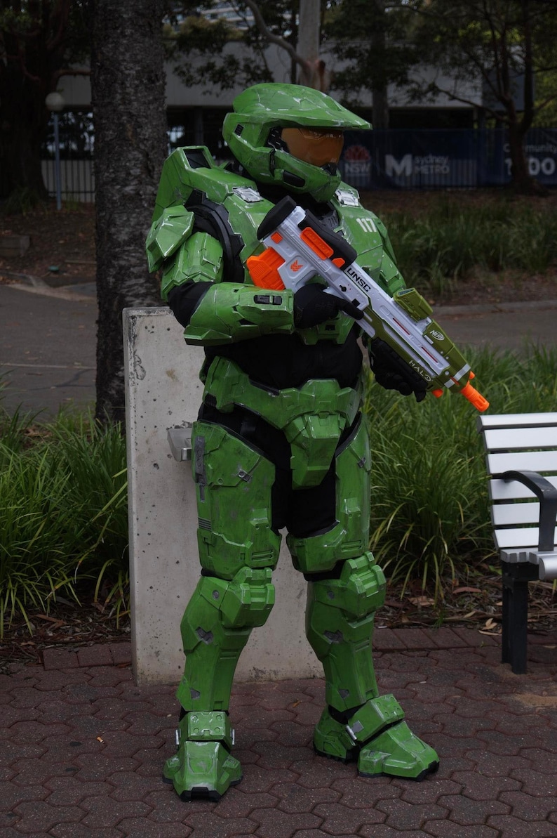 May include: A green and black  'Halo'  video game character costume with a white toy gun. The gun has orange accents and the text 'UNSC' and 'HALO' printed on it. The character is wearing a green helmet with a visor.