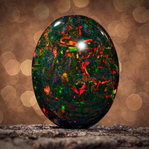 May include: An oval-shaped black opal gemstone featuring a dark base color with vibrant, multi-colored flashes of red, green, and blue throughout its surface. The stone has a smooth, polished finish with a high-gloss sheen reflecting light.