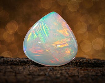 May include: A pear-shaped, iridescent opal gemstone with a milky white base and flashes of green, blue, and red. The gemstone sits on a dark, textured surface, with a blurred, golden bokeh background.
