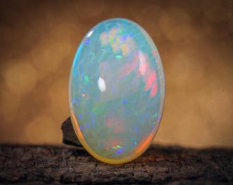 May include: A large, oval opal gemstone with a milky white base and iridescent flashes of color. The gemstone is set against a blurred, warm-toned background, and rests on a piece of dark wood. The opal displays a play of color.