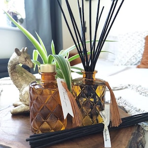 May include: Two amber glass bottles with black diffuser sticks and brown tassels. The bottles have a diamond pattern and are filled with a clear liquid. The bottles are on a wooden surface with a small gold giraffe figurine. The tag on the bottle reads "Thank you" and "Handmade".