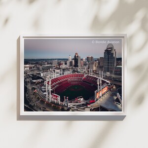 Pode incluir: Uma vista aérea do Great American Ball Park, casa dos Cincinnati Reds, com o horizonte da cidade ao fundo. O estádio é principalmente vermelho com um campo verde e linhas brancas. O horizonte da cidade é uma mistura de edifícios altos e edifícios mais baixos. O céu é azul claro com algumas nuvens.