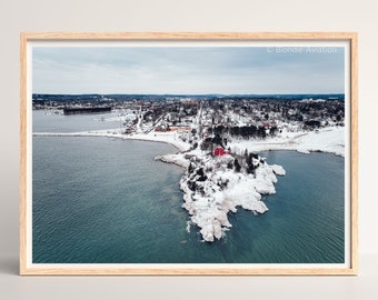 May include: An aerial view of a small town on a snowy lakeshore. A red house sits on a rocky outcrop with snow-covered trees. The water is a deep blue and the sky is cloudy.