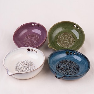 May include: Four ceramic grater bowls in purple, green, white, and blue. Each round bowl features a textured, flower-shaped center for grating, a small pouring spout on the rim, and three small circular holes along the edge for hanging or storage.