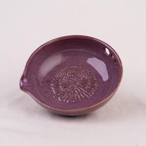 May include: A round, purple ceramic grater bowl with a glossy finish. The interior base features a raised, circular, starburst-like texture for grating. The rim includes a small pouring spout on one side and three small circular holes on the other.