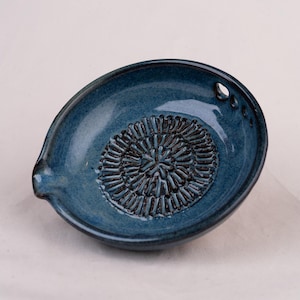 Può includere: Un piatto grattugia rotondo in ceramica blu con un motivo radiale scuro e strutturato al centro per grattugiare il cibo. Il piatto presenta un piccolo beccuccio su un lato e un foro circolare vicino al bordo per appenderlo, con smalto lucido.