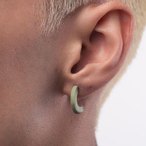 Könnte beinhalten: Nahaufnahme eines hellgrünen Creolen-Ohrrings. Der Ohrring hat ein gebogenes, C-förmiges Design. Der Ohrring wird am Ohr einer Person mit kurzen, blonden Haaren getragen.