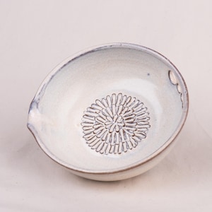 May include: A round, off-white ceramic bowl with a textured, dark brown floral pattern embossed in the center. The bowl features a small pouring spout on the left side and a single circular hole near the rim on the right side, with a smooth glaze.
