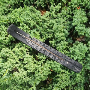 May include: A black wooden incense holder with carved runes and a groove for the incense stick. The holder is rectangular with a slightly rounded end. The background is a bed of green foliage.