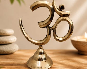 May include: A gold-colored Om symbol statue. The symbol is mounted on a decorative base. The statue is displayed on a wooden surface, with a lit candle and a stack of stones in the background. The Om symbol is a spiritual icon.