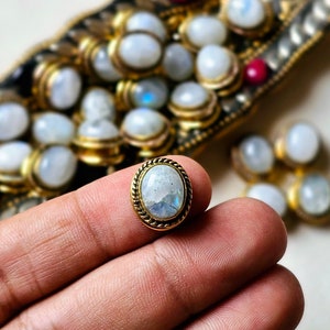 Può includere: Un cabochon ovale di pietra di luna incastonato in una lunetta di metallo dorato con un bordo a forma di corda. La pietra di luna ha un colore bianco latte con riflessi blu e iridescenti. Altri cabochon di pietra di luna sono visibili sullo sfondo.