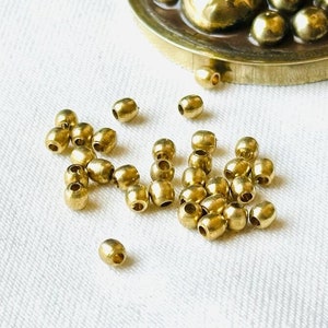 以下が含まれることがあります： 白い布の表面に散らばった、金色の金属ビーズの山。ビーズは楕円形で、滑らかで光沢のある仕上がりです。