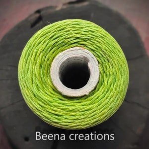 Puede incluir: Una bobina de hilo verde brillante enrollada alrededor de un tubo de cartón blanco. El hilo está enrollado apretado y tiene un ligero brillo. El texto "Beena creations" es visible en la parte inferior de la imagen.