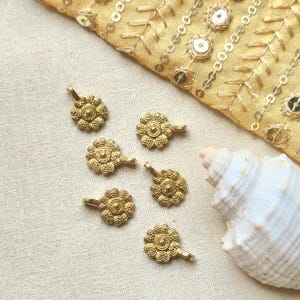 May include: Six gold-colored, flower-shaped pendants with small loops. The pendants are arranged on a cream-colored fabric, with a gold sequined fabric and a white seashell in the background.