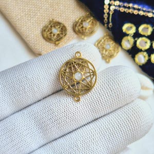 May include: Gold-toned metal charm with a starburst design and a white gemstone in the center. The charm has a loop on each side for attaching to jewelry.