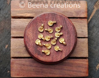 May include: A close-up of a group of 16 gold-colored metal charms, each with a small loop at the top. The charms are arranged on a wooden surface.