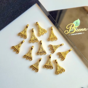 Puede incluir: Una colección de hallazgos de joyería dorados, cada uno con un diseño texturizado en forma de abanico y múltiples bucles para sujetar a otros componentes. Los hallazgos están dispersos sobre una superficie blanca, con una etiqueta negra y dorada en el fondo.
