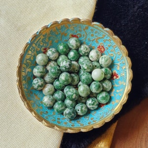 Puede incluir: Un cuenco lleno de cuentas de ágata verde y blanca. Las cuentas son redondas y tienen un patrón moteado. El cuenco está decorado con un diseño floral azul y dorado.