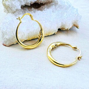 May include: A pair of gold-colored hoop earrings. Each earring features a crescent moon design with a face carved into the moon. The earrings are displayed on a light-colored surface with a white crystal formation in the background.